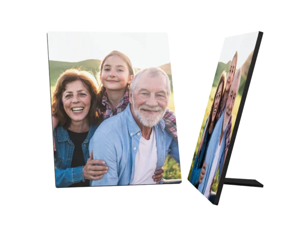 Ein Fotoaufsteller zeigt ein fröhliches Familienbild. Eine lächelnde ältere Frau mit braunen Haaren steht links. Hinter ihr steht ein junges Mädchen mit blonden Haaren in einem karierten Hemd und lächelt ebenfalls. Rechts von ihnen steht ein älterer Mann mit weißen Haaren und Bart, der auch fröhlich in die Kamera schaut und seine Arme um die beiden anderen gelegt hat.